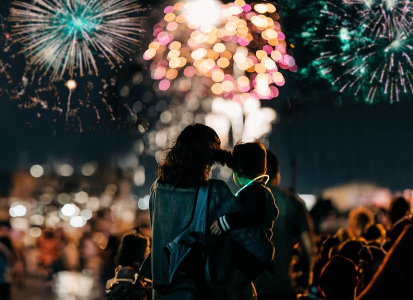 Mother and child facing with their backs to the camera, watching a colourful fireworks display ahead of them.