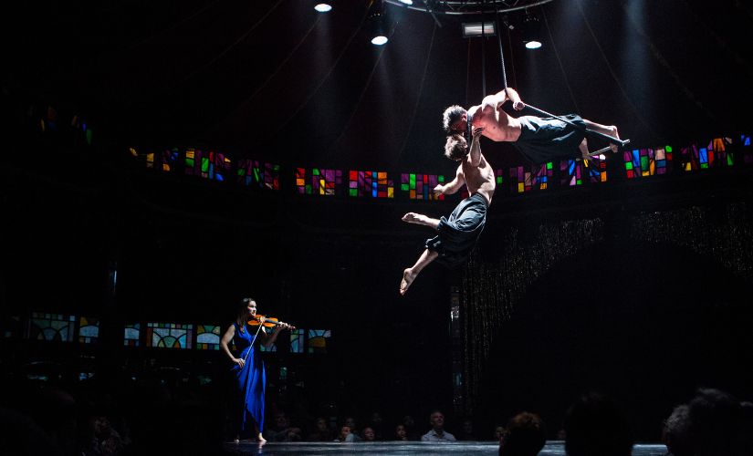 Two circus performers hanging from a trapeze accompanied by a violinist