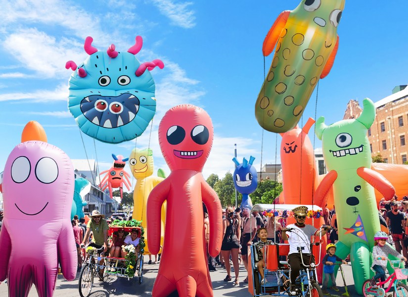 A crowd participating in the Big Bike Parade, riding bikes and scooters alongside giant inflatable cartoon creatures.