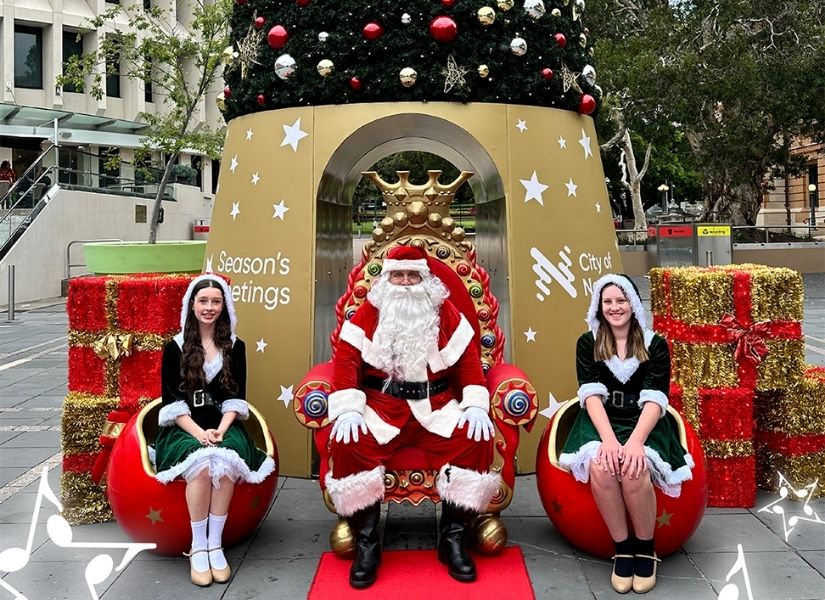 Santa sitting on a red throne in front of a giant Christmas tree in Wheeler Place, with two of Santa's helpers sitting either side.